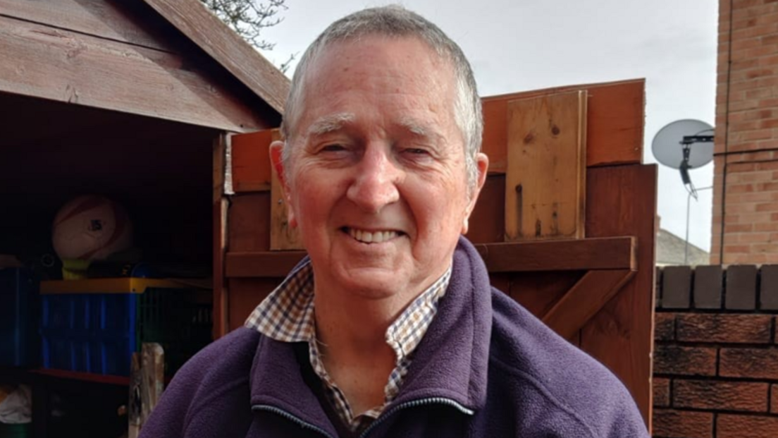 David Watch wearing a chequered shirt underneath a purple three-quarter-zip fleece jumper. He is clean shaven with short grey hair and is smiling at the camera, standing in a garden beside a wooden shed with the door open.