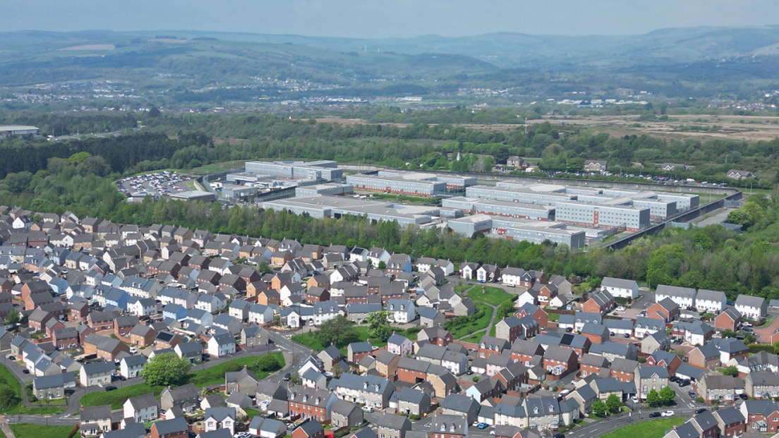 A drone image of Parc Prison, in front of it is the Parc Derwen housing estate can be seen, with hills in the distance behind it 