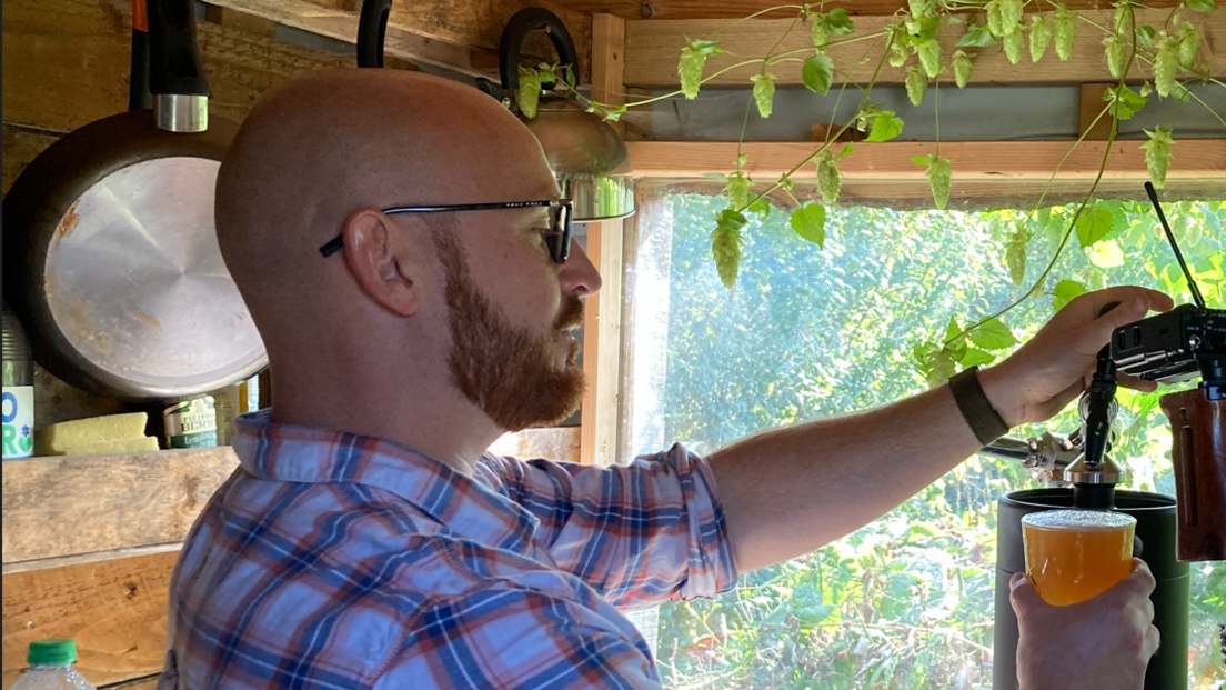 A bald man with a beard, wearing a checked shirt pulling a pint in a shed