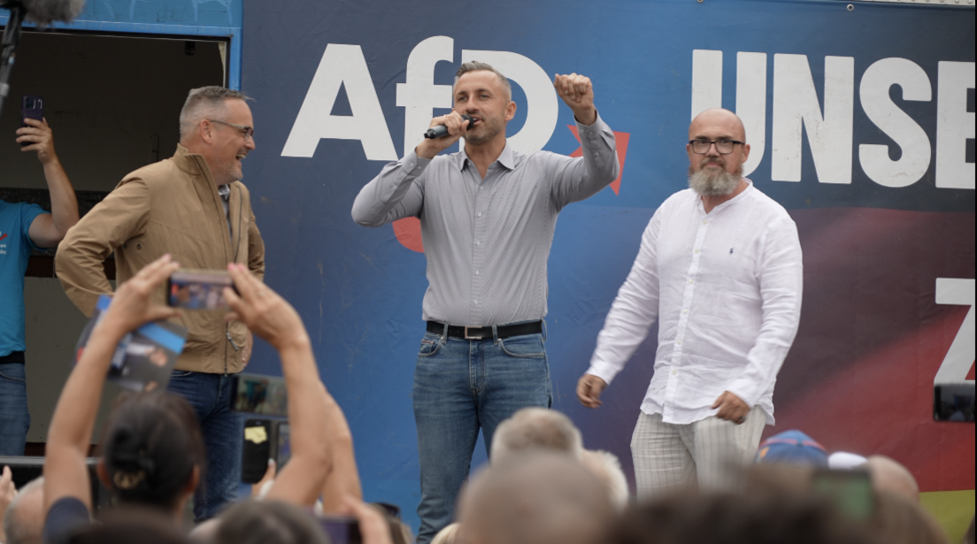Three men are on-stage astatine an AfD rally. Ulrich Siegmund is successful the centre holding a microphone. Crowds are listening and filming connected their phones.