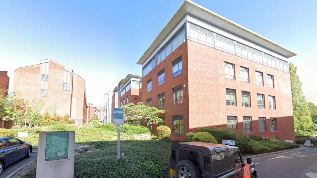 An brick office block with a white flat roof called Senate House, part of the Southernhay Gardens development in Exeter. A jeep-style car is parked on the road in front with an orange hi-vis jacket hanging off the wing mirror. There are green bushes and trees surrounding the building.