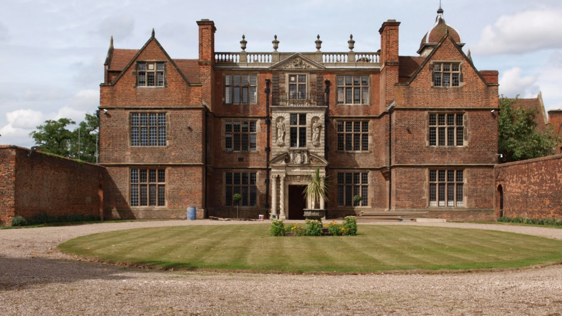 The grand building stands behind a circular lawn and flora. It has five sections with windows on three levels and an ornate frontage and entrance.