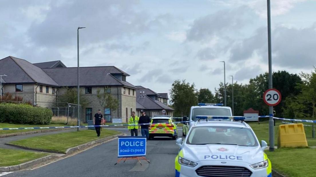 Police cars in front of a cordon, a sign reads Police Road Closed, you can see the hospital building in the background.
