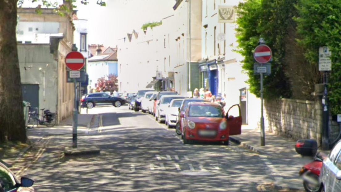 Two no entry signs sit on either side of the end of a one-way road. The road has cars parked along one side of it and has one lane for vehicles to drive along.