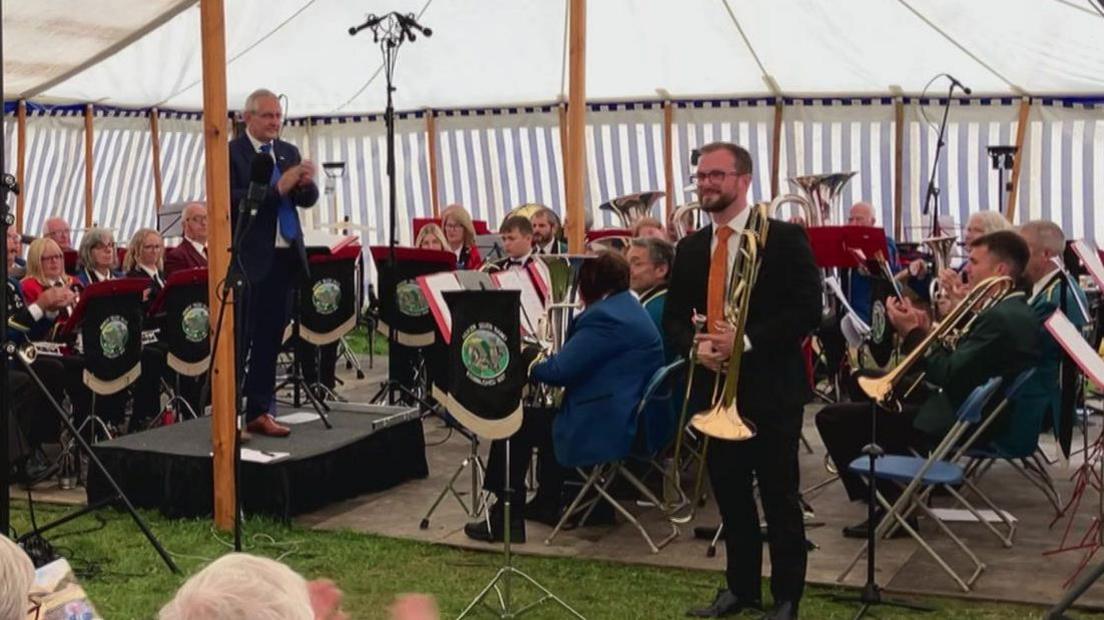 A group of musicians wearing brightly coloured blazers and ties hold brass instruments, sat at the front of a marquee in a performance formation. A 30-year-old man with an orange tie stands in front as the soloist, holding a trombone. A man in his 50s stands on the conductor's podium applauding the soloist, his son.