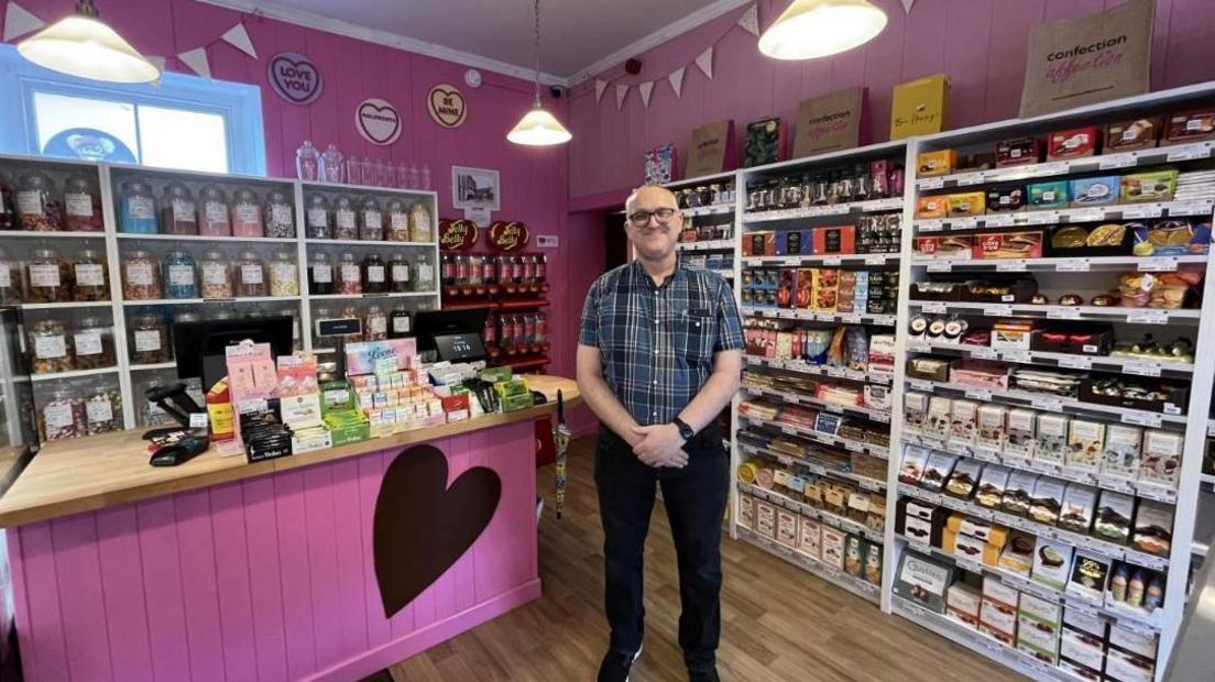 The image shows the interior of a cheerful sweet shop with pink-painted walls and a playful, festive atmosphere. Shelves are stocked with a variety of confectionery, and a wooden-topped counter featuring a large heart design on the front displays boxes of sweets, jars, and other treats. Behind the counter, glass jars are filled with assorted sweets, and a Jelly Belly jelly bean display is visible on the right-hand side. Decorative touches such as bunting and circular signs reading "LOVE YOU," "HAPPY BIRTHDAY," and "BE MINE" hang from the ceiling, adding to the celebratory feel. A person wearing a checked shirt and black trousers stands in front of the counter with their hands clasped together.