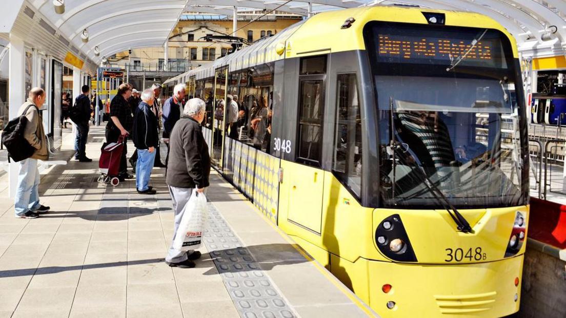 People boarding a Metrolink tram at Manchester Victoria station on a sunny day. The tram information display shows 'Bury via Victoria',