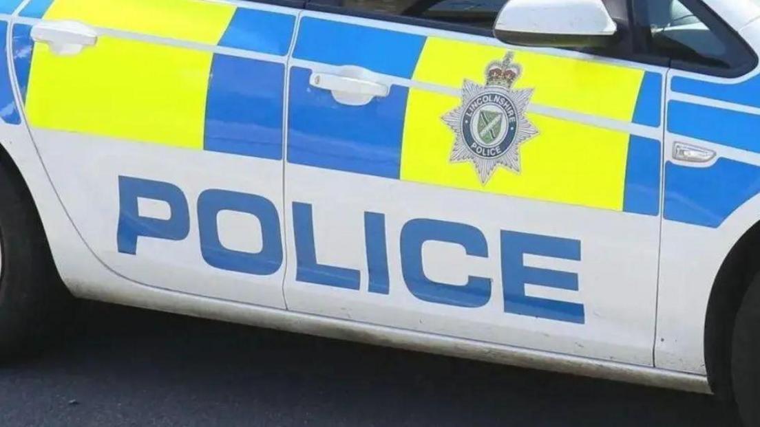 Side view of a marked Lincolnshire Police car. It is white with yellow and blue blocks and the force's badge on the door.