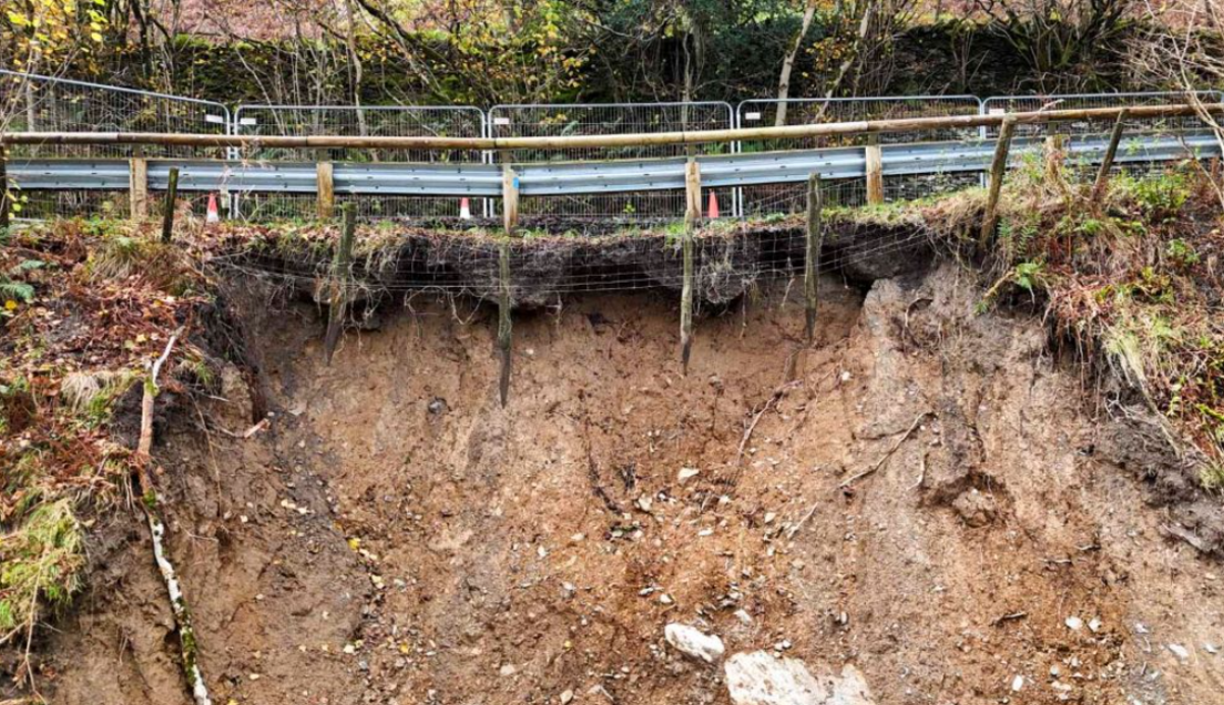 A grey fence can seen above the soil of the landslip. The bank is steep and rocks can be seen jutting out of the orange-tinged soil. Orange cones are on the road. 