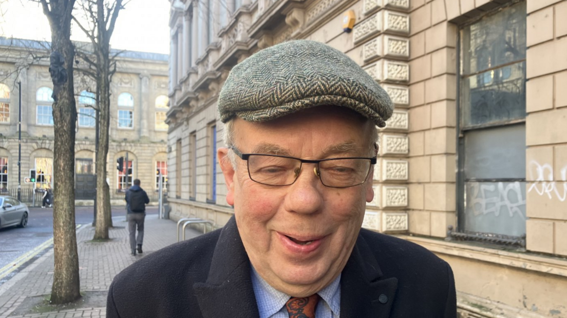 John Gray, wearing a cap, glasses, dark jacket, blue shirt and brown tie