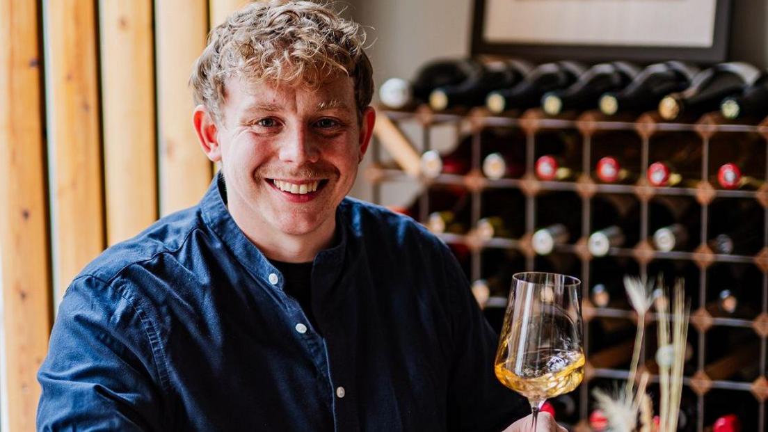 Joseph Stark, who has created a non-alcoholic drinks menu holds a wine glass with an amber-coloured drink in it. A wine rack stands behind him containing rows of bottles. Mr Stark is wearing a blue shirt and smiling at the camera, looking happy.