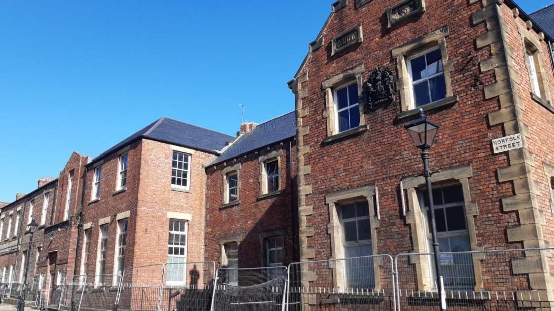 An imposing two-storey red brick building with the Norfolk Street sign. There is metal railing in front of it.
