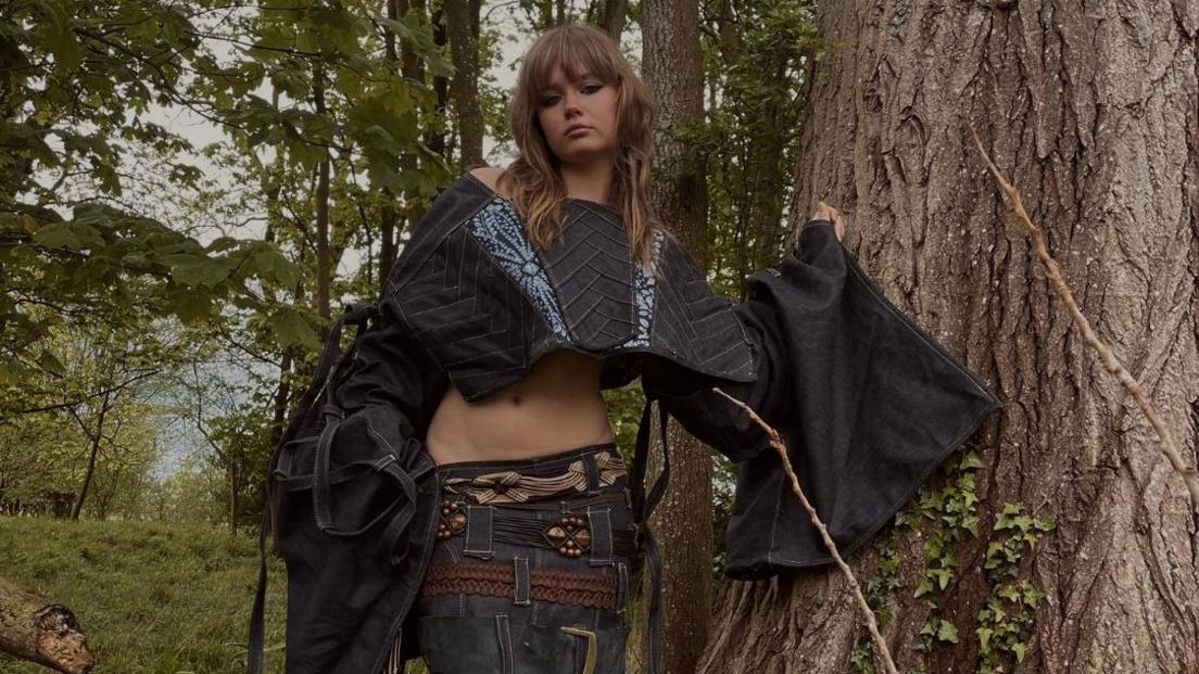 Alexandra Burch wearing one of her garments - a two piece denim-looking top with oversized sleeves and a skirt. She is in the woods, by a tree.