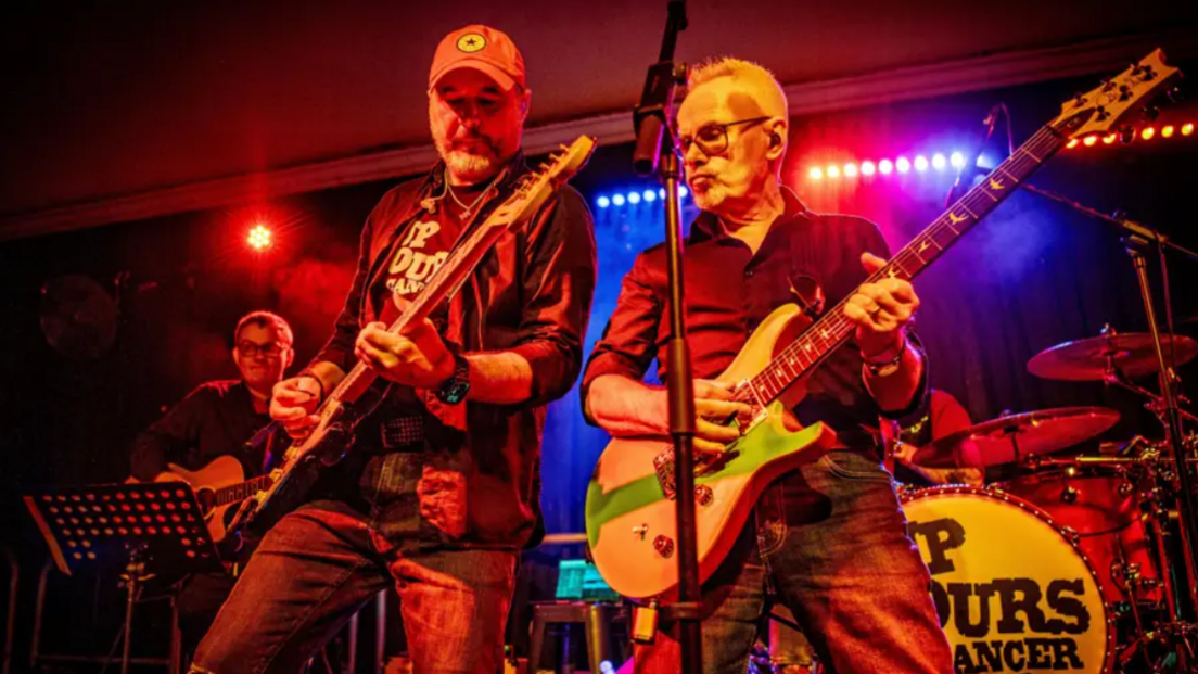 Two men are standing with guitars on a stage, with a microphone in the middle of them. In the background is another man with a guitar, and a pair of drums. 