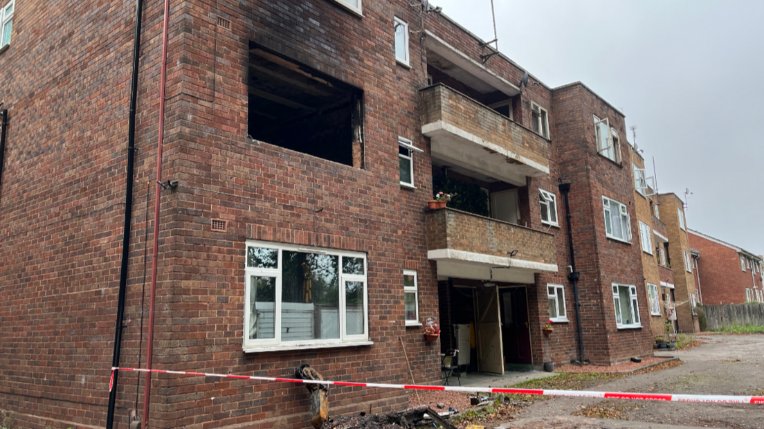 A three-storey brick building, with a second floor window missing, and scorch marks up the wall. Red and white fire service tape stretches out from a pipe at ground level, with burnt debris behind it