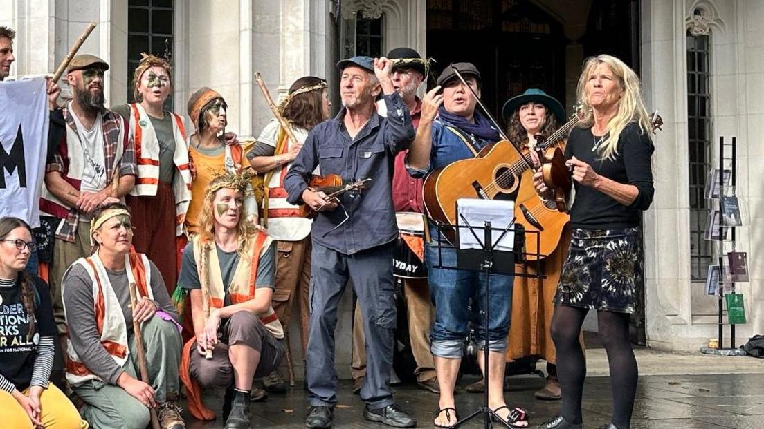 Three musicians, one holding a guitar and two holding violins join campaigners outside the Supreme Court in London, with some campaigners wearing heavy make-up and floral headwear.