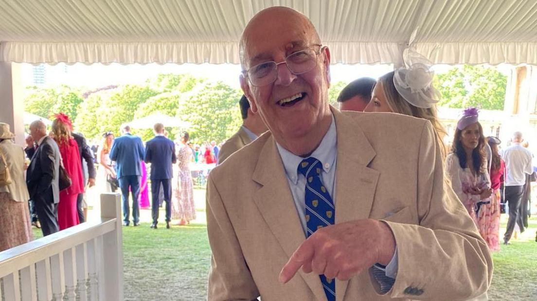 A man is seen smiling directly at the camera. He is wearing a beige suit with a blue striped tie. He has silver thin glasses and is seen to be pointed at something off camera.