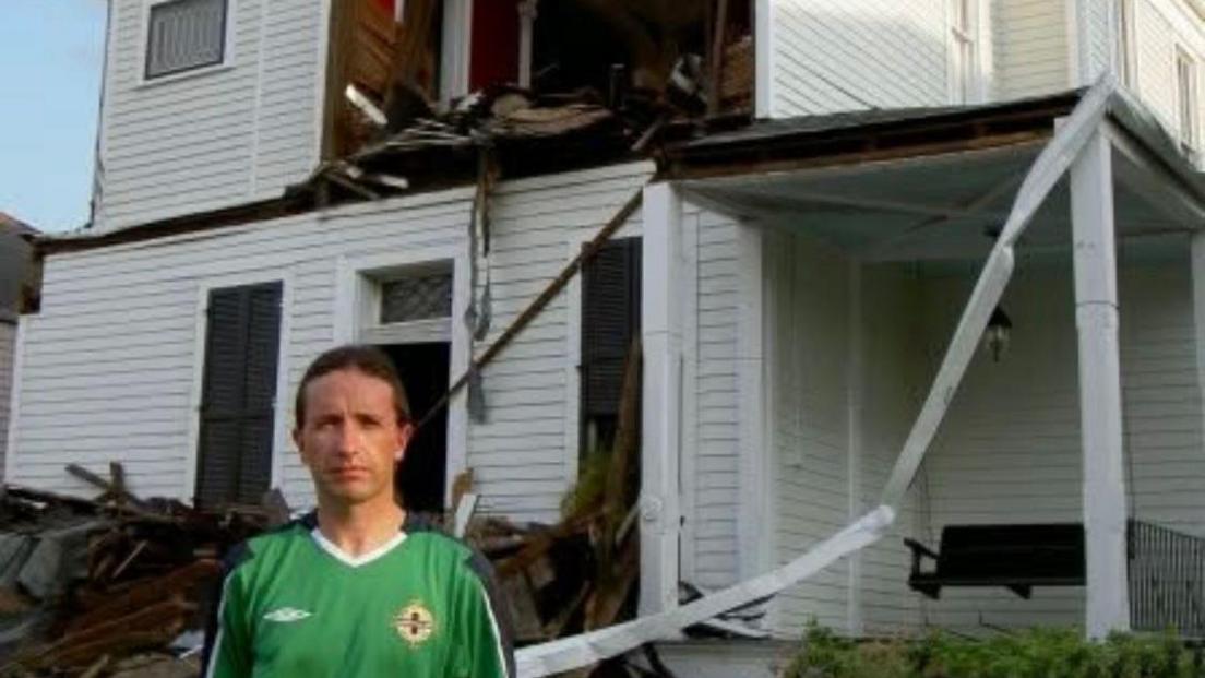 Stephen Rea, with short  brown hair, looks at the camera. He is wearing a green Northern Ireland football jersey which is green with dark blue trim. Behind him is a badly damaged white clapboard house - a section of the wall on the first floor has collapsed and brown wooden debris is strewn in a heap behind Stephen Rea.