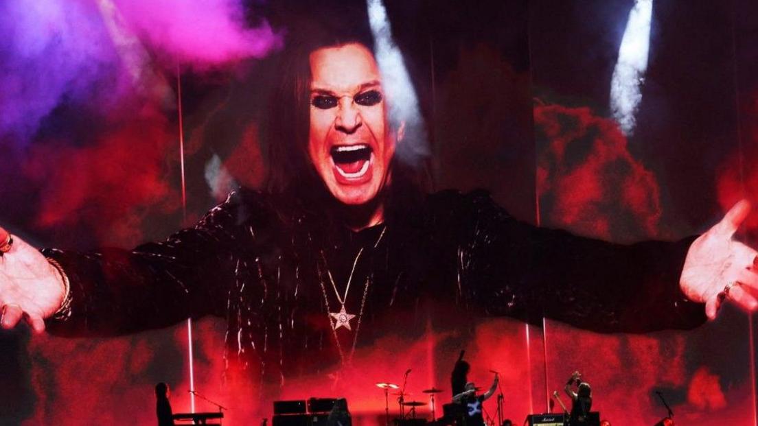 An huge picture of Ozzy Osbourne, with his mouth wide open in his trademark snarl, wearing black and his arms outstretched. The image is bathed in smoke and red and purple lights above musicians performing on stage.