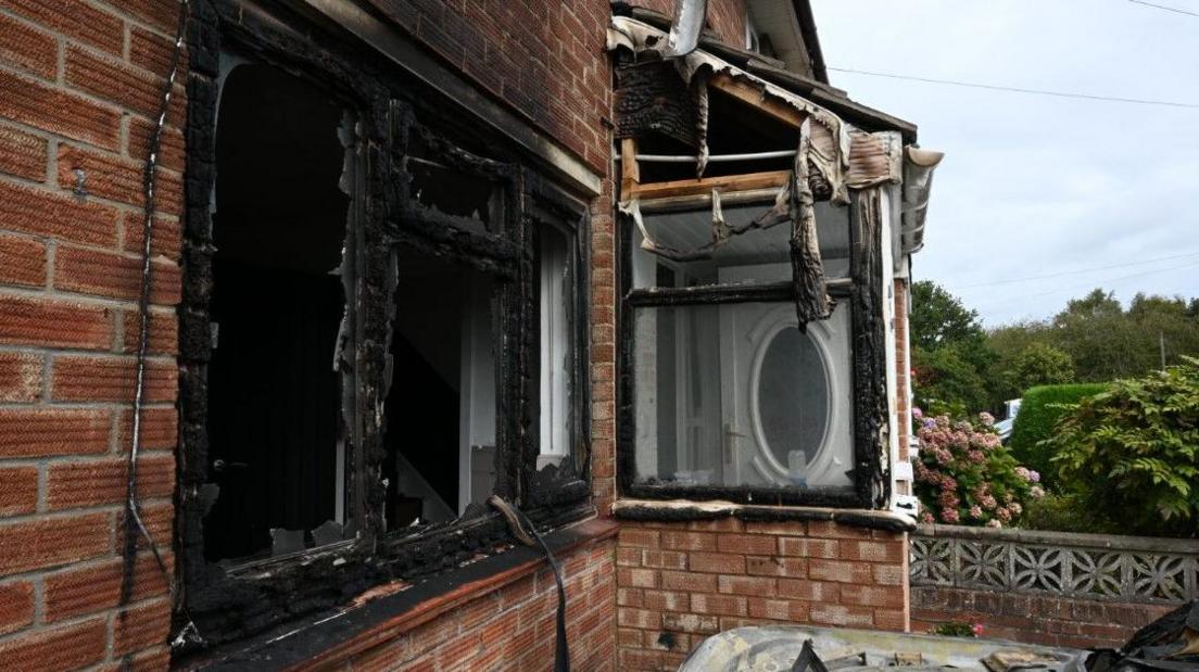 A red brick property with its windows and doors charred black. The bonnet of a burnt-out car is just visible in the foreground.