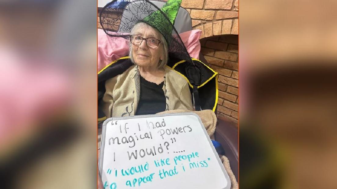 An elderly woman sitting in a chair with a pink pillow wearing glasses and a witches hat holding up a sign that says "if I had magical powers I would?" with the answer "I would like people to appear that I miss".