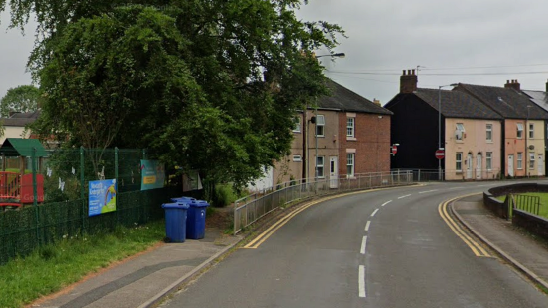 A road, bending to the right, with a children's playground behind a fence on the left of the road, followed by terraced houses 