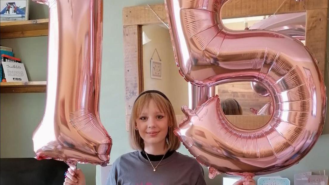 A teenage girl with shoulder length blonde hair wearing a silver necklace and grey jumper smiling into the camera. She is stood in a room of the house holding up two pink balloons, one says 1 and the other says 5.