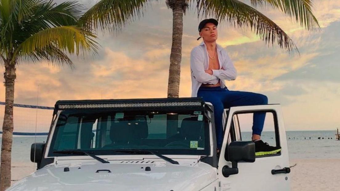 Samy Daim sitting on top of a white 4x4 vehicle parked beside a beach with  palm trees in the background. Mr Daim is wearing an unbuttoned white shirt with blue trousers and cap.