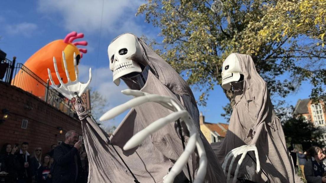 Two skeleton figures walking through the streets of Lincoln. They are wearing grey capes. An orange inflatable monster is in the back left of the photo. 