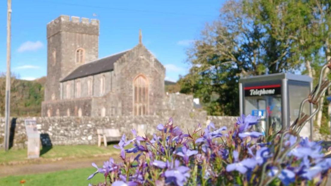 A simple stone-built church showing nave and tower. There is a stone wall in front of the church, with trees to the right of the building. There is a modern telephone kiosk near the camera, and some bluebells in the foreground.