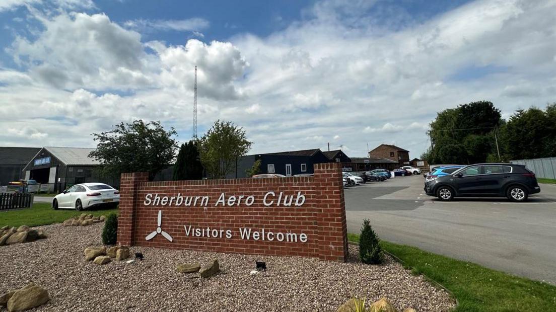 Sherburn Aero Club entrance, showing a brick wall with a sign attached and a car park behind