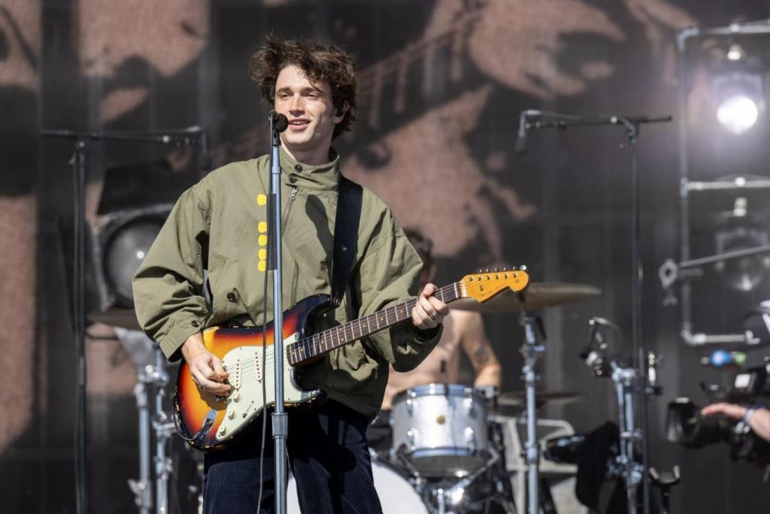 A man with a fender stratocaster and a green jacket in front of a mic stand with five yellow plectrums