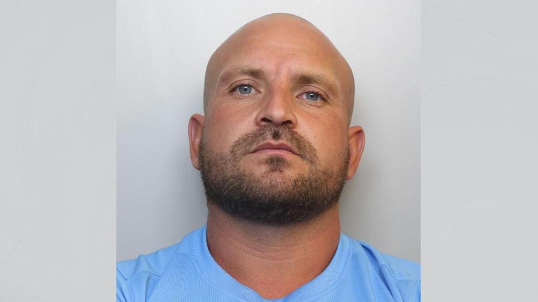 A police mugshot of Ashley Hunt. He is bald with a dark brown beard and a neutral facial expression. He is wearing a light blue t-shirt and standing against a grey background.