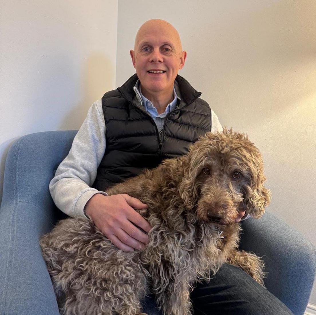 Gary Tait sits in a light blue fabric chair against a plain cream wall. He is wearing a blue shirt, grey jumper and a padded gilet. A big shaggy light brown dog sits on his knee and Gary has a hand across its curly fur.