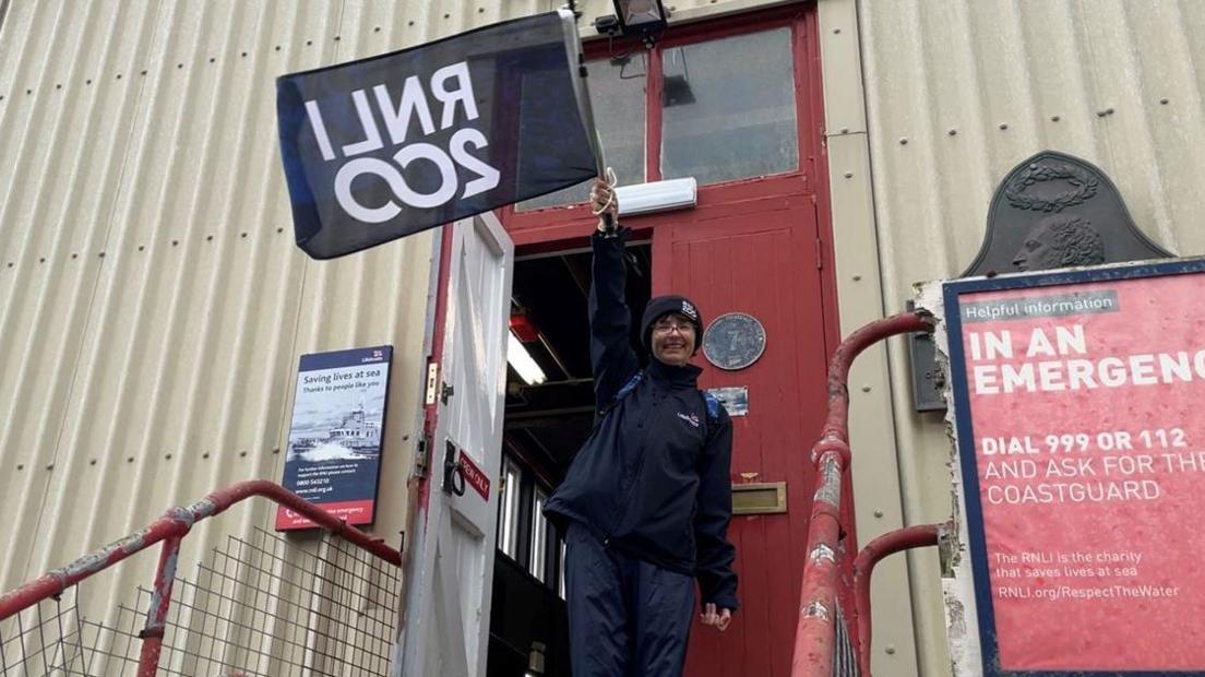 Anjie Rook dressed in navy blue waterproofs and hat standing outside the doorway of the Douglas Lifeboat station waving a flag navy blue flag. The phrase RNLI 200 can be seen in back to front lettering on the flag.