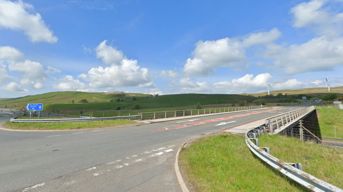 Junction 37. It is a two-lane road with a blue sign which reads 'The South M6'. There are cars in the distance and two wind turbines on the hills to the right.
