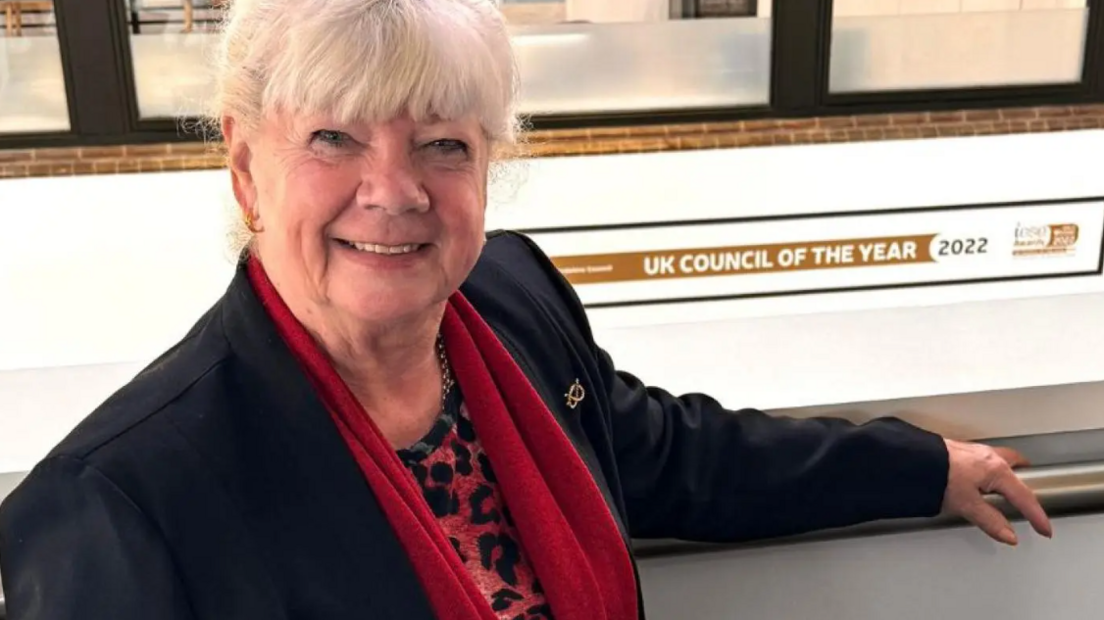 A woman with light grey hair, a black jacket and red scarf smiles at the camera, with one arm leaning on a rail