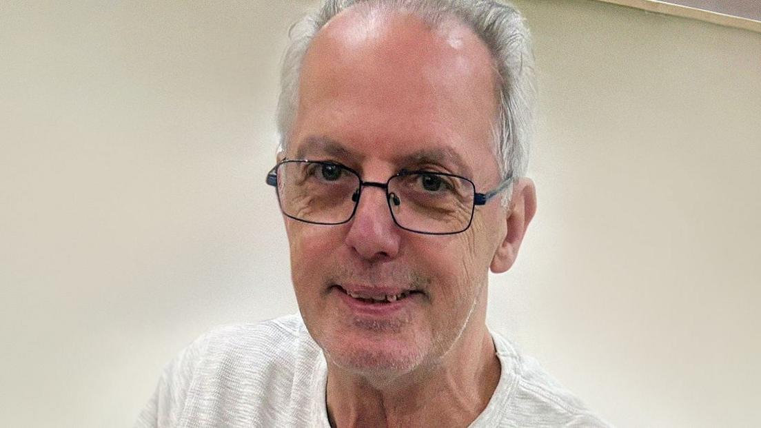 Man in glasses with grey hair balding at front looks at camera with slight smile, head and shoulders shot wearing white t shirt.