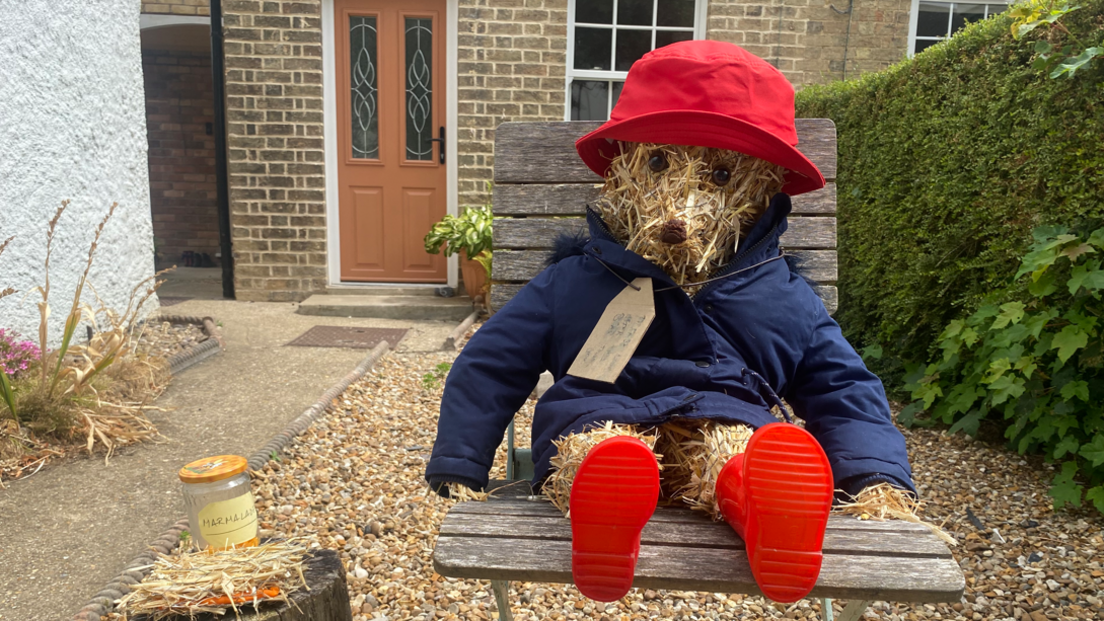 A Paddington Bear scarecrow sits on a chair outside a house. It has a red hat, red boots and a blue coat with a label tied around its neck. Next to it is a jar of marmalade. 