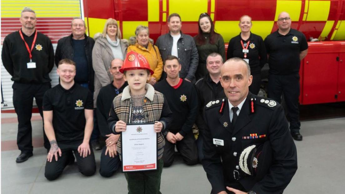 A four-year-old boy is stood next to Chief Fire Officer Rob Barber who is wearing his ceremonial uniform and holding a cap under his arm. There are a group of firefighters and others stood behind them, in front of a fire service water tanker.