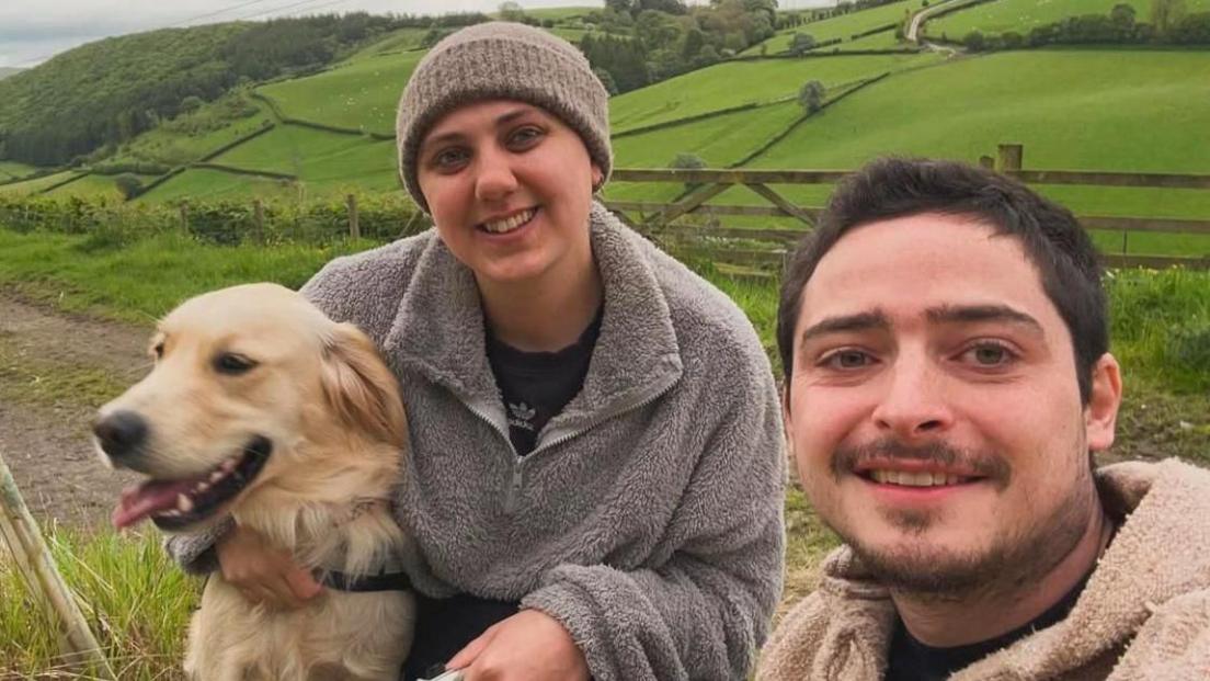 A photo of a woman in the middle cuddling a golden retriever. She is smiling and wearing a grey hat and a grey fleece. A man is to the right of her and has dark hair as well as a moustache and beard and has a cream-coloured fleece on. There are fields behind them. The man is taking the selfie.