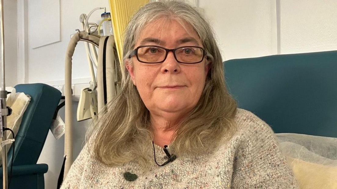 Lesley Jenkins, who has long grey hair and glasses, sat on a hospital chair with a cubical curtain and medical equipment behind her. She is wearing a grey jumper with a broadcast mic pinned to it.