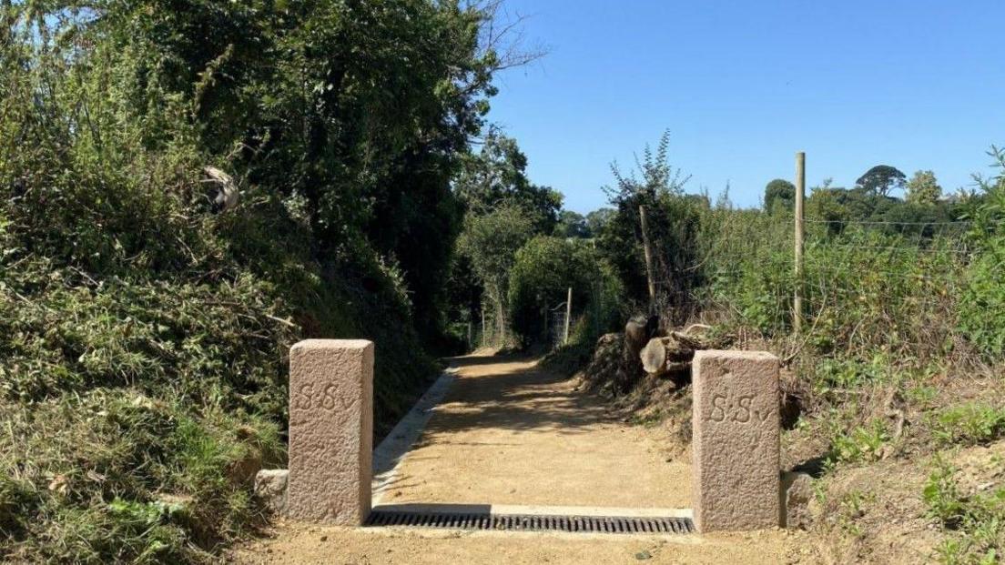 Path in St Saviour at Val Aumé, with two stone pillars at the entrance and greenery either side 