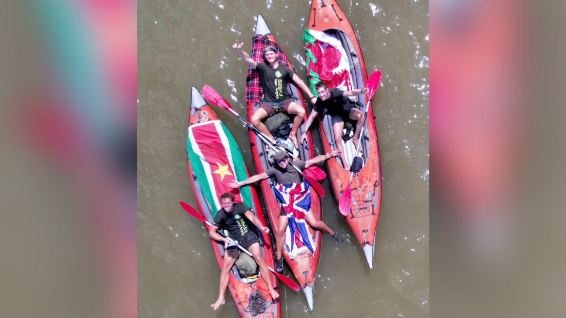 Ash Dykes and team members  Dick Lock, Matt Wallace and Jacob Hudson are photographed from above lying on three of the four kayaks, smiling. The Suriname and Welsh flags and Union Jack and a Scottish tartan are draped across the kayaks. The water looks brown and murky.