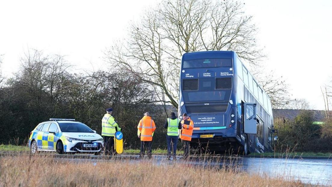 A door is open at the back of the bus with the front of the vehicle in a roadside ditch. A police car is nearby with officers at the scene. There is a bollard in the road.