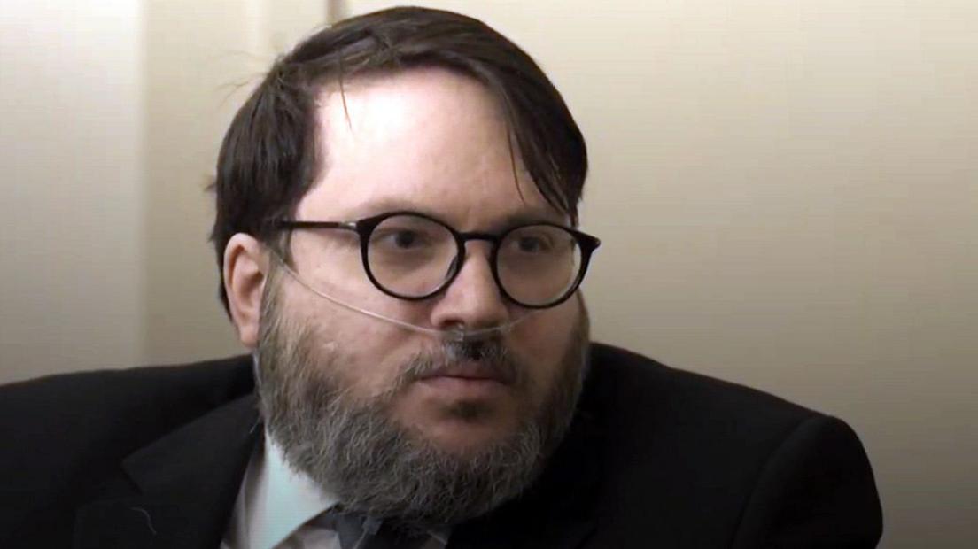 A bearded man with an oxygen tube in his nose. He is wearing glasses and a dark suit
