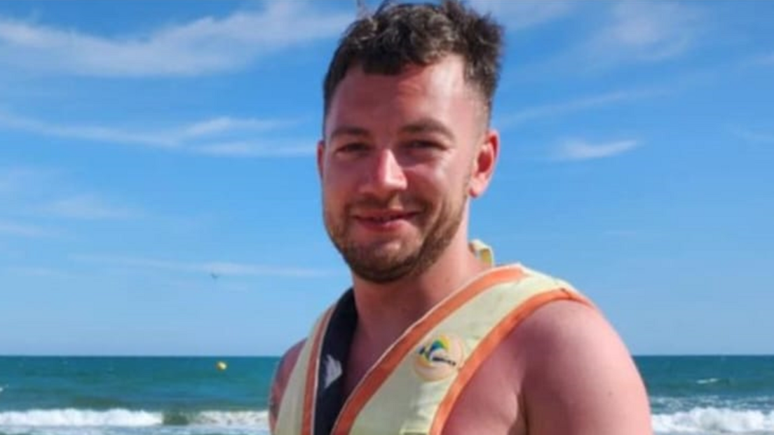 Kris, wearing a life jacket or harness, smiles on a beach with breaking waves behind him