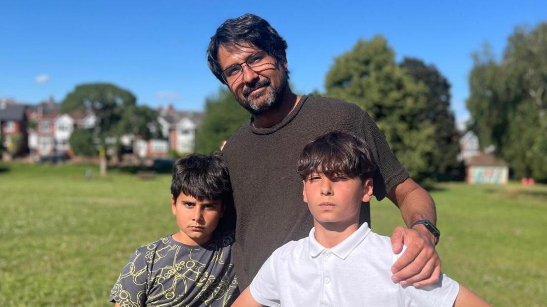 Hugo in a dark olive T-shirt with his arms around his sons Luca, in a grey and yellow patterned T-shirt, and Guilherme in a white polo shirt, in a park in Exeter.