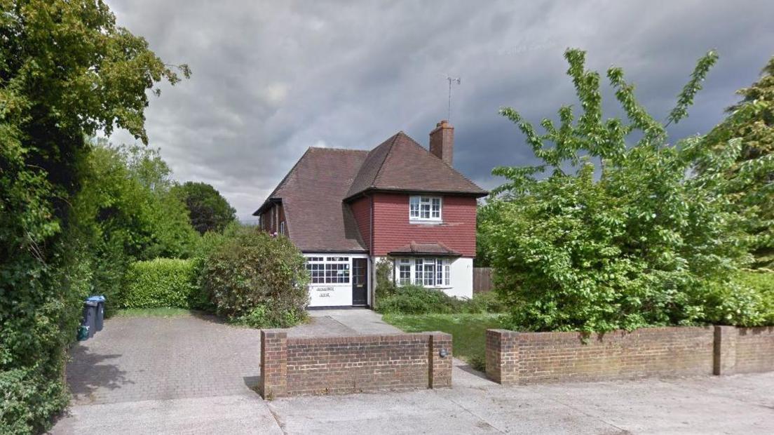 The outside of a detached house which has a white rendered lower half and red tiled about. There is a driveway and a front lawn with shrubs growing around the edge as well as trees with green leaves behind a low brick wall