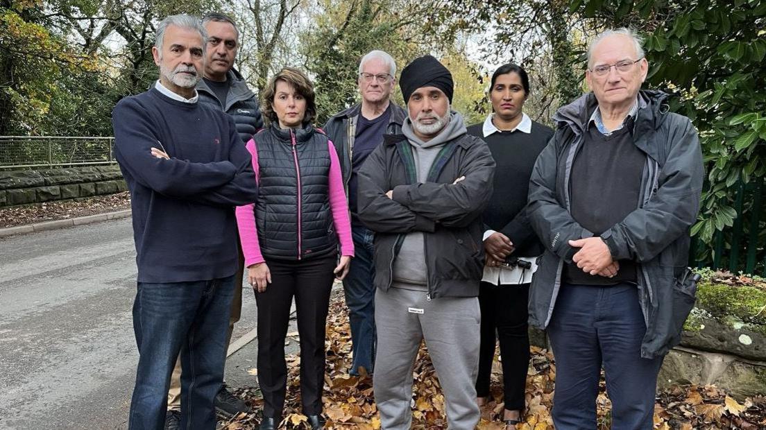 A group of residents standing on a road, with no pavement, looking at the camera. There are trees and autumn leaves on the ground